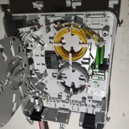 Otwarta skrzynka telekomunikacyjna z widocznymi kablami światłowodowymi w kolorach żółtym, zielonym i transparentnym, zamontowana na szarej ścianie.