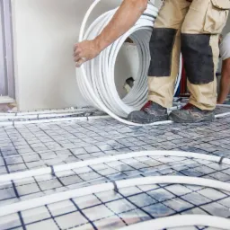 Instalacja ogrzewania podłogowego: białe rury układane na folii z siatką, monter w roboczych spodniach i butach trzyma zwój rur, drugi monter w tle pracuje nad instalacją.