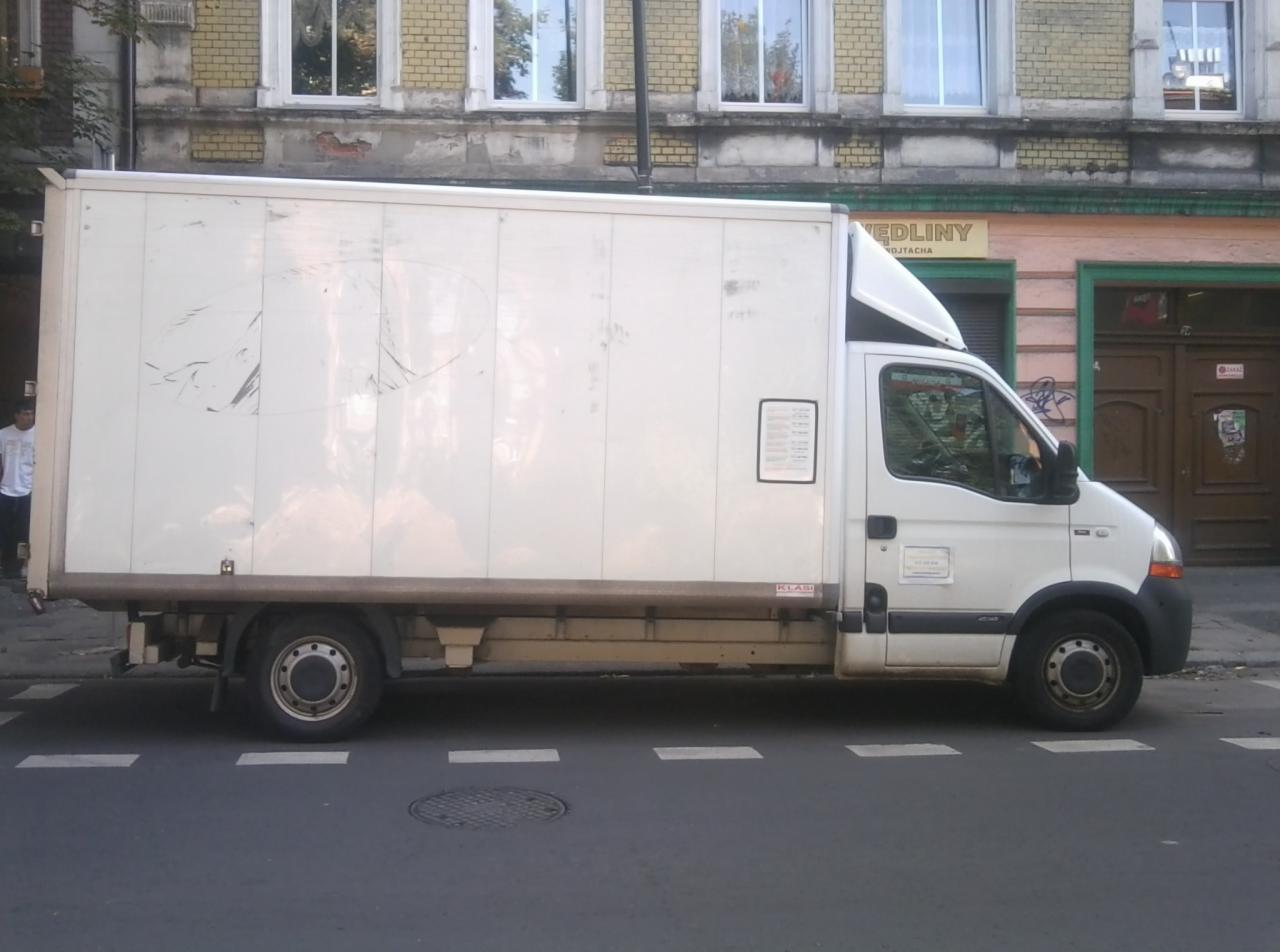 Biały bus dostawczy zaparkowany na ulicy z widocznymi śladami użytkowania na karoserii, na tle kamienicy z oknami i szyldem sklepu. Widok z boku.