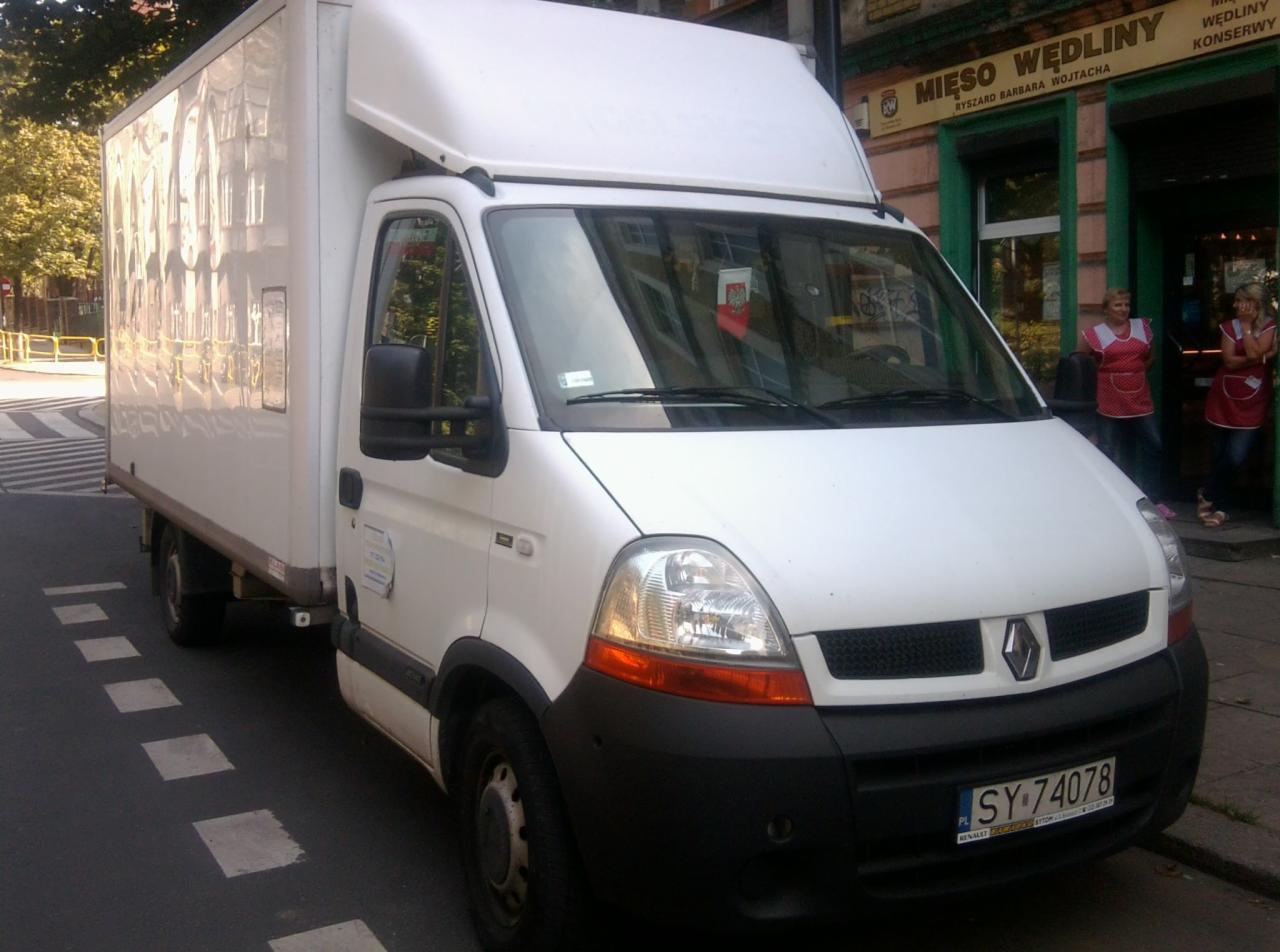 Biały Renault Master z zabudową kontenerową zaparkowany na ulicy, na tle sklepu mięsnego z personelem w fartuchach. Rejestracja SY 74078.