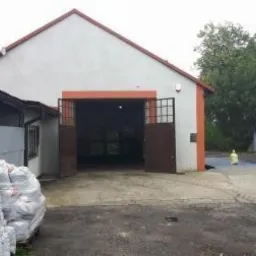 Składowisko ekogroszku: białe worki z nadrukiem, ułożone przed szarym budynkiem magazynowym z otwartą bramą i stertą węgla w tle.