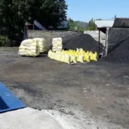 Składowisko z hałdami czarnego węgla i paletami worków z żółtymi i białymi opakowaniami, widoczne betonowe boksy i zieleń w tle.