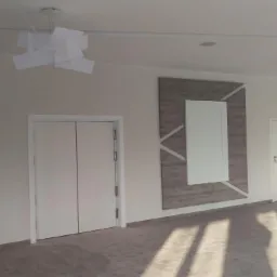 Wykończone wnętrze przestronnego pomieszczenia z dwiema parami białych drzwi, dekoracyjnymi panelami ściennymi w odcieniach szarości i białymi, nowoczesnymi lampami sufitowymi.