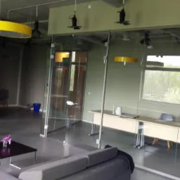 Nowoczesne wnętrze biurowe z szarymi meblami wypoczynkowymi, szklanymi ścianami działowymi, okrągłymi lampami w kolorach zielonym i ż&oacute;łtym oraz reflektorami sufitowymi.