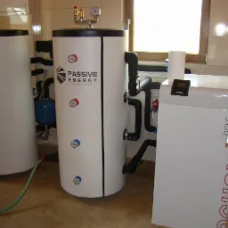 Instalacja wewnętrzna z dwoma zbiornikami akumulacyjnymi ciepła, rurami i armaturą, oraz jednostką sterującą Ochsner Golf, zainstalowana w pomieszczeniu z jasnymi płytkami ceramicznymi.