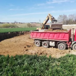 Żółta koparka ładuje ziemię do czerwonej wywrotki podczas prac ziemnych na polu, widoczny wykop i zielona trawa na pierwszym planie.