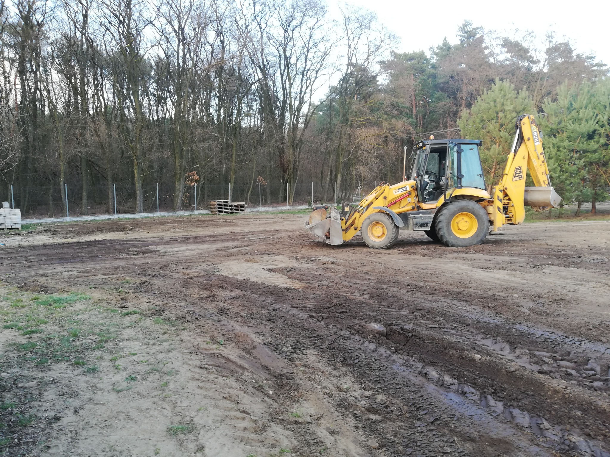 Żółta koparko-ładowarka JCB na placu budowy z widocznymi śladami opon w błocie, w tle las i palety z kostką brukową.
