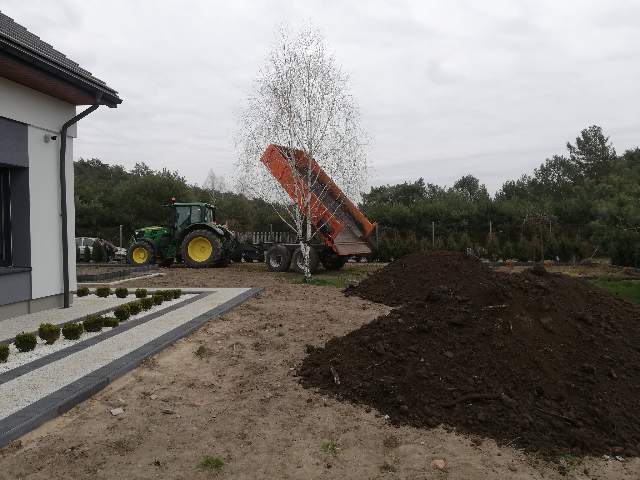 Pomarańczowa wywrotka podnosi skrzynię, wysypując ziemię obok świeżo wybudowanego domu z zagospodarowanym terenem i traktorem w tle.