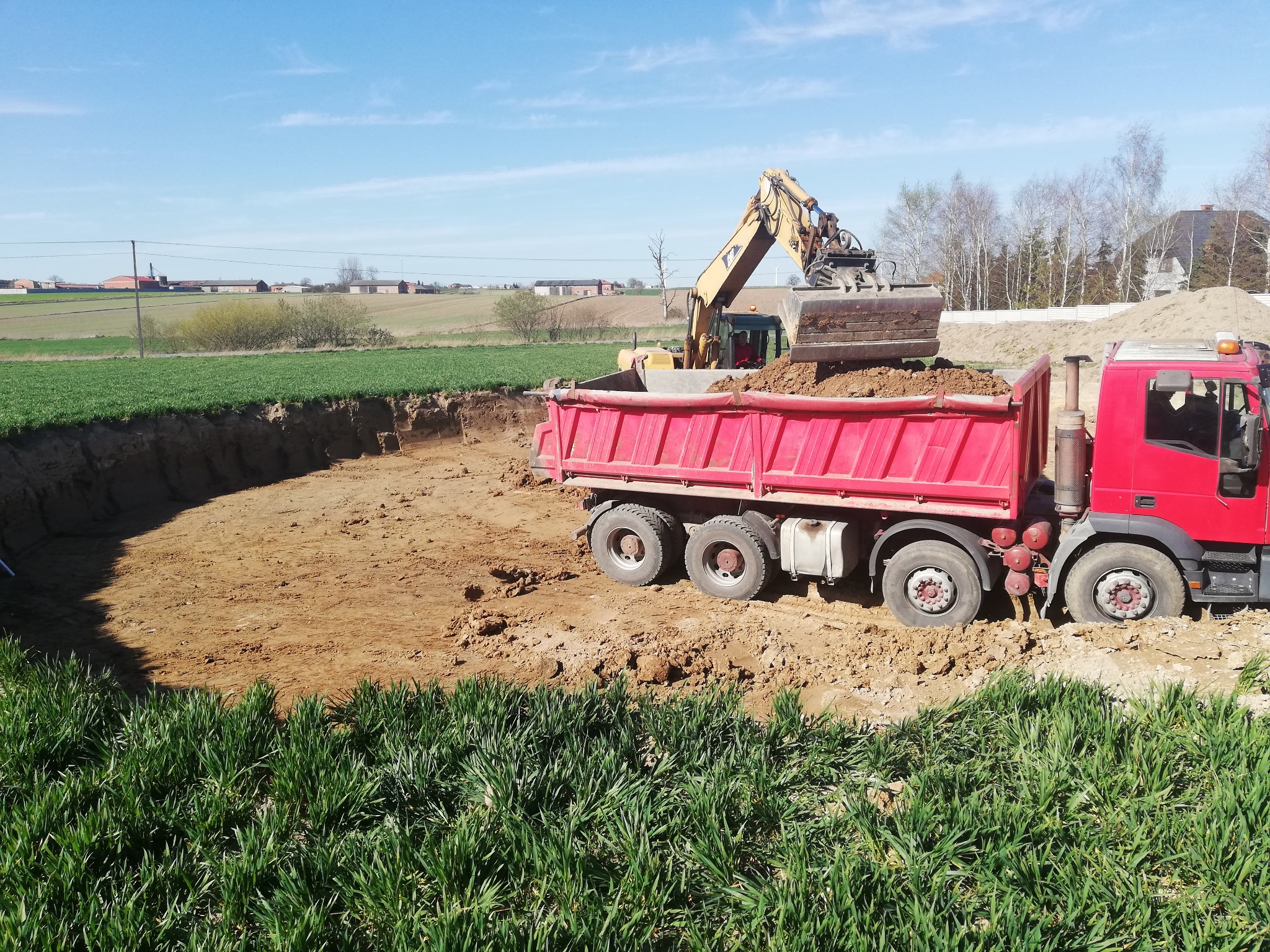 Żółta koparka ładuje ziemię do czerwonej wywrotki podczas prac ziemnych na polu, widoczny wykop i zielona trawa na pierwszym planie.