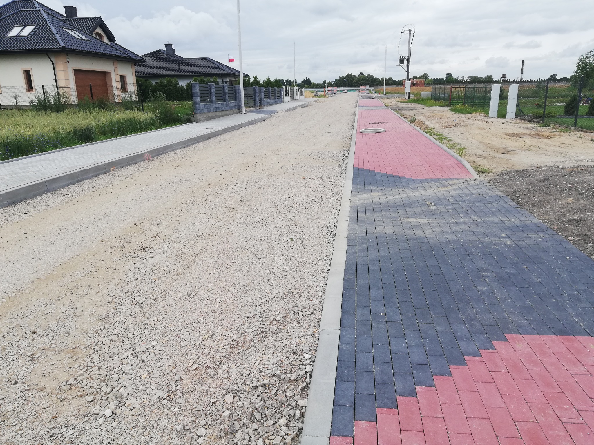 Ulica w trakcie budowy z częściowo ułożoną kostką brukową w kolorach czerwonym i szarym, widoczny podsypka z kruszywa, w tle domy jednorodzinne i ogrodzenia.