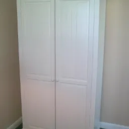 Biała, dwudrzwiowa szafa z dekoracyjnym gzymsem, umieszczona na tle beżowej ściany i ciemnej podłogi. Prosty, klasyczny design.