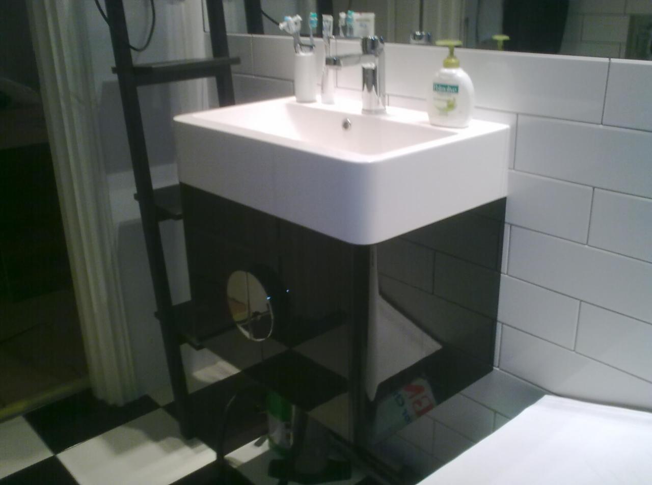 Nowoczesna umywalka nablatowa z czarną szafką i lustrem w łazience z białą cegłą na ścianach i czarno-białą szachownicą na podłodze.