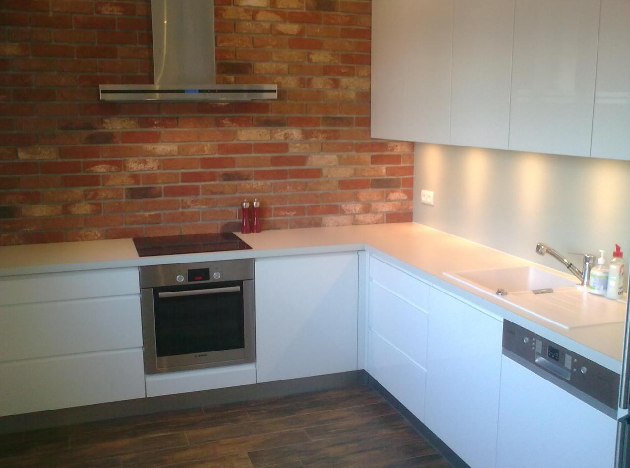 Nowoczesna kuchnia z białymi szafkami i ceglaną ścianą. Widoczny piekarnik, płyta indukcyjna, okap i zlew. Uwagę przyciąga kontrast materiałów i oświetlenie.