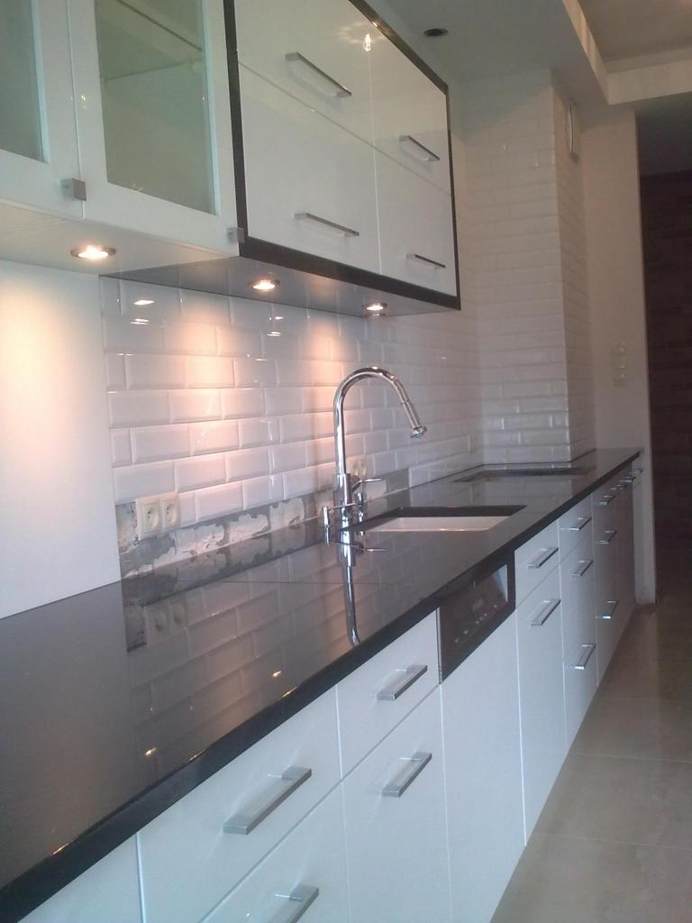 Biała kuchnia z czarnym blatem i białymi płytkami typu metro. Bateria kuchenna chromowana. Szafki górne z przeszklonymi frontami. Oświetlenie punktowe pod szafkami.