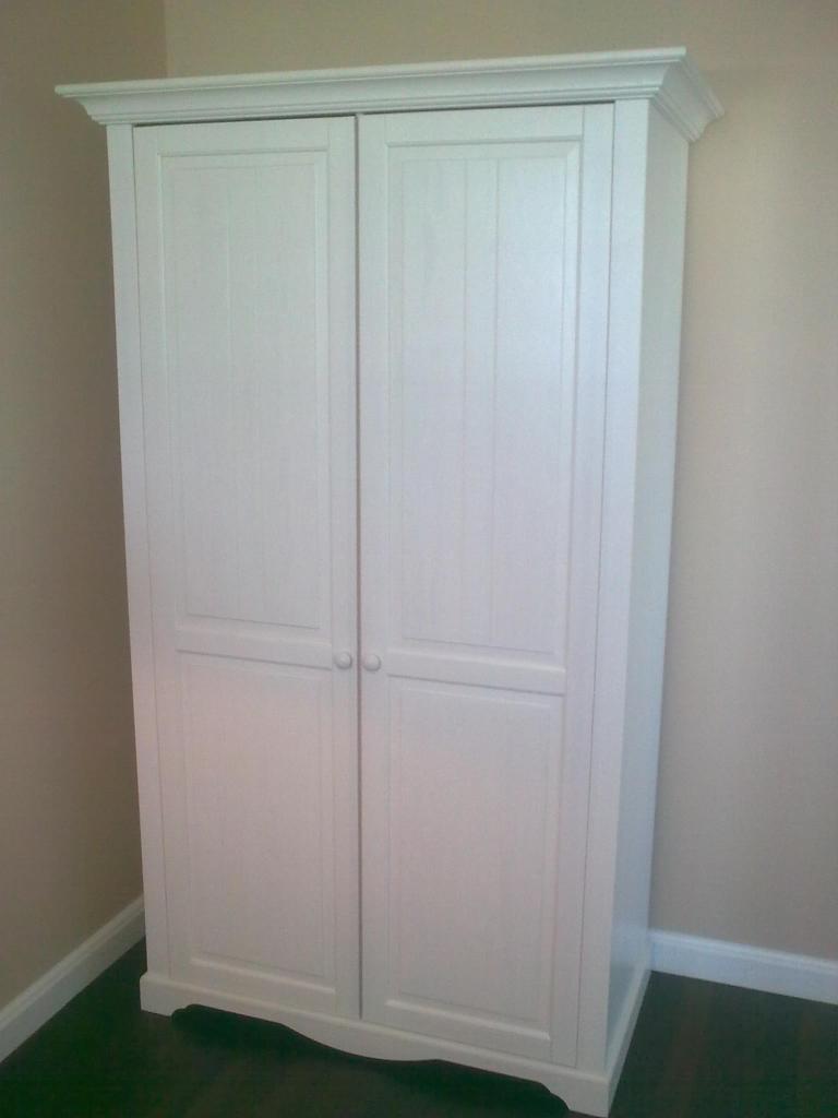 Biała, dwudrzwiowa szafa z dekoracyjnym gzymsem, umieszczona na tle beżowej ściany i ciemnej podłogi. Prosty, klasyczny design.