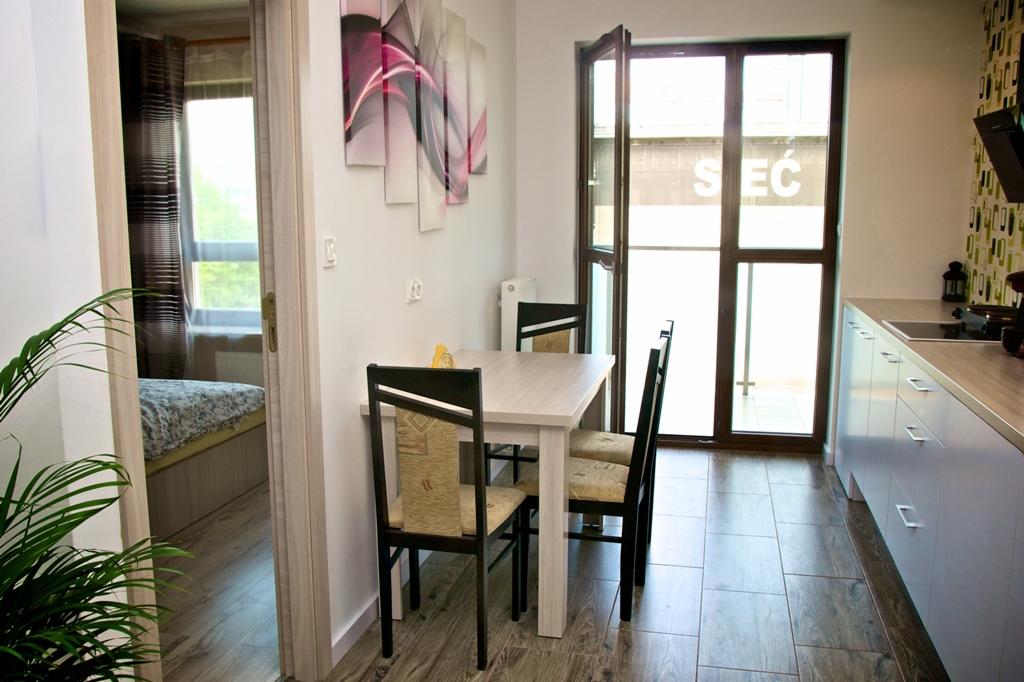 Jasna kuchnia z jadalnią: stół z krzesłami, zabudowa kuchenna z blatem, okno balkonowe. W tle widoczny pokój z łóżkiem i dekoracje ścienne.