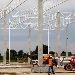 ROSA-HALE Sp. z o.o. - Wznoszenie hali: stalowa konstrukcja szkieletowa z betowymi słupami, na budowie pracownik w pomarańczowej kamizelce i maszyny budowlane na tle zieleni.