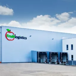 Nowoczesna hala magazynowa Fruitlogistics z rampami załadunkowymi i biurowcem, błękitne niebo z chmurami w tle. Elewacja z paneli w kolorze jasnoszarym.