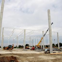 ROSA-HALE Sp. z o.o. - Budowa hali: betonowe słupy, stalowa konstrukcja dachu, żuraw i koparki na placu budowy. Widoczne zbrojenia i elementy szalunków.