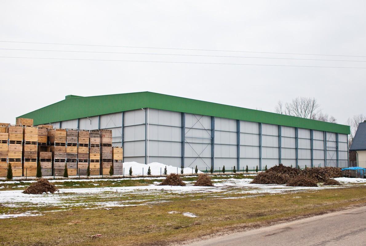 Zimowy krajobraz z dużą, zielono-białą halą magazynową o konstrukcji stalowej, skrzyniami i resztkami śniegu na polu.