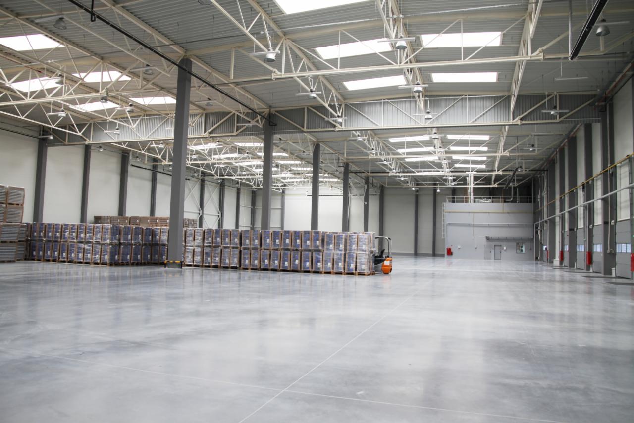 Wnętrze hali magazynowej z metalową konstrukcją dachu i rzędami zapakowanych palet. Na posadzce widoczny wózek widłowy. Oświetlenie sufitowe.