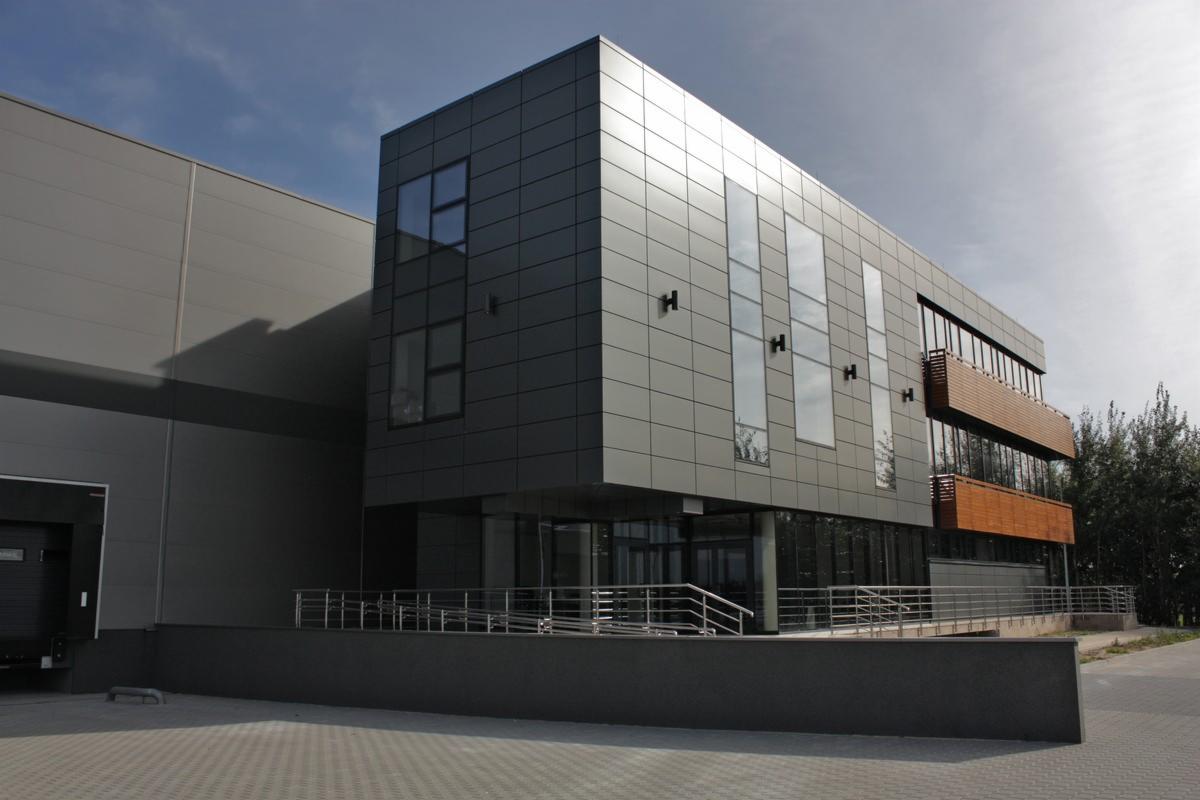 Nowoczesny budynek przemysłowy z szarymi panelami i drewnianymi balkonami, widoczny z zewnątrz, w słoneczny dzień. Szklane panele w fasadzie.