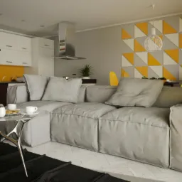 Nowoczesne wnętrze: szary narożnik z poduszkami, designerski stolik kawowy, kuchnia w tle z żółtym akcentem, geometryczna dekoracja ścienna w odcieniach żółci i bieli.