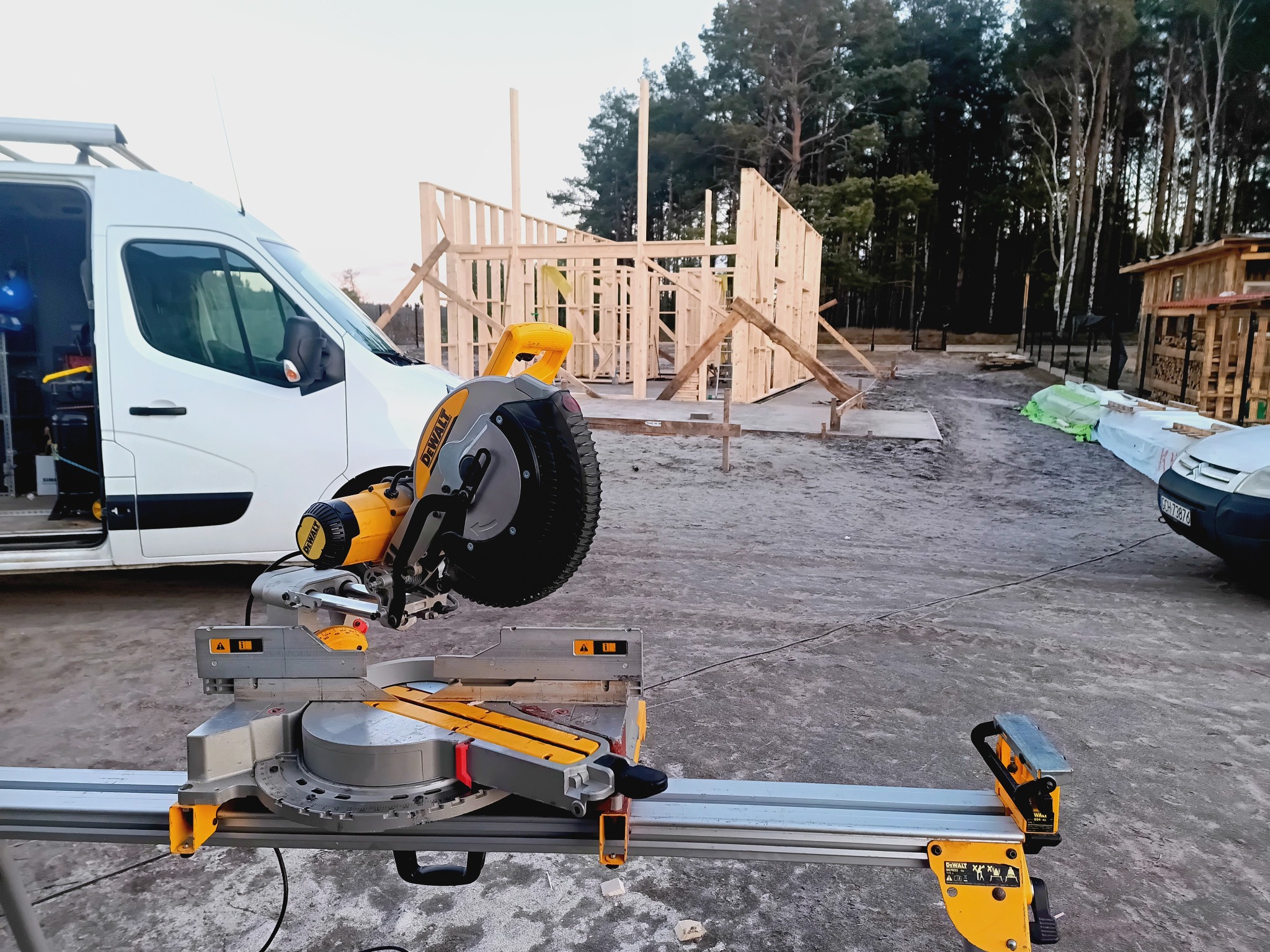 Budowa domu szkieletowego w toku: widoczna konstrukcja i piła DeWalt na tle lasu i białego busa. Etap wznoszenia ścian, narzędzie gotowe do pracy.