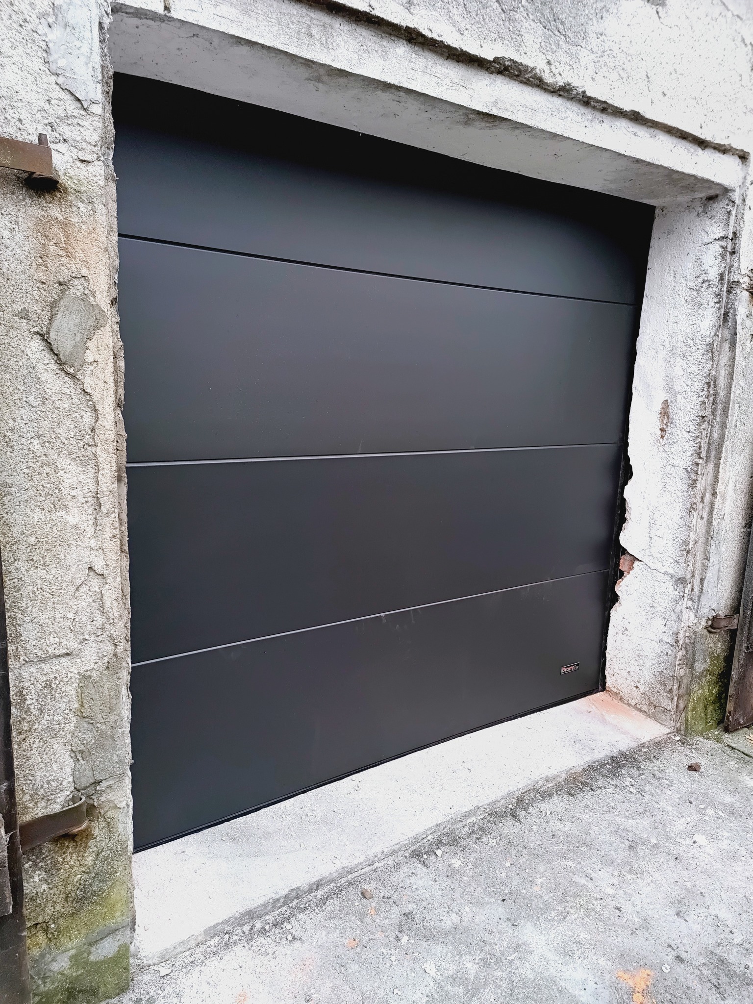 Zamknięta, segmentowa brama garażowa w kolorze antracytu, z widocznym fragmentem zniszczonego muru wokół otworu wjazdowego. Minimalistyczny design bramy.