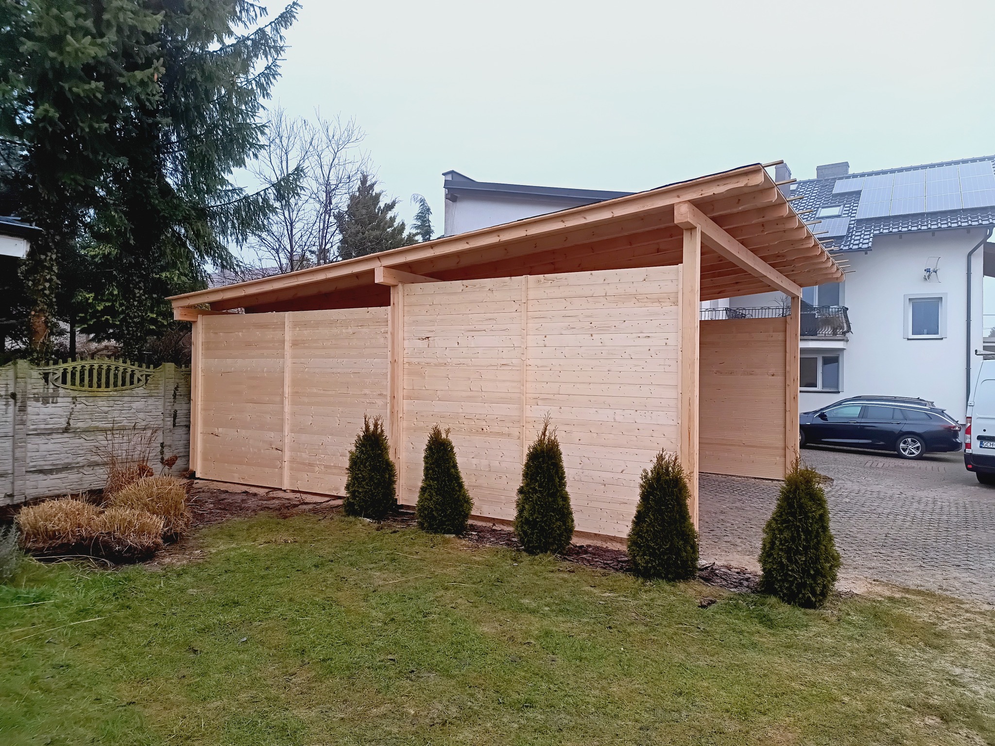 Drewniana wiata garażowa z płaskim dachem, obok domu jednorodzinnego. Wzdłuż ściany wiązki posadzone tuje. Na podjeździe zaparkowany samochód.