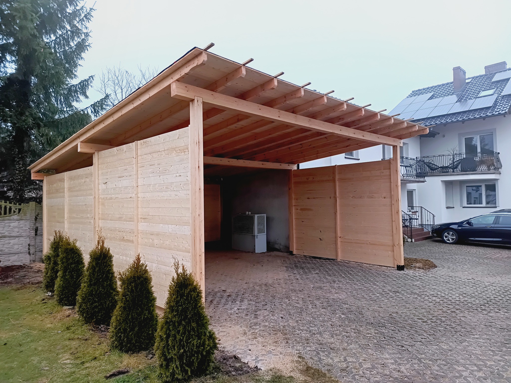 Drewniana wiata garażowa z płaskim dachem i częściowym obłożeniem ścianami z desek, z widocznym wyposażeniem wewnątrz, na tle domu z panelami słonecznymi.