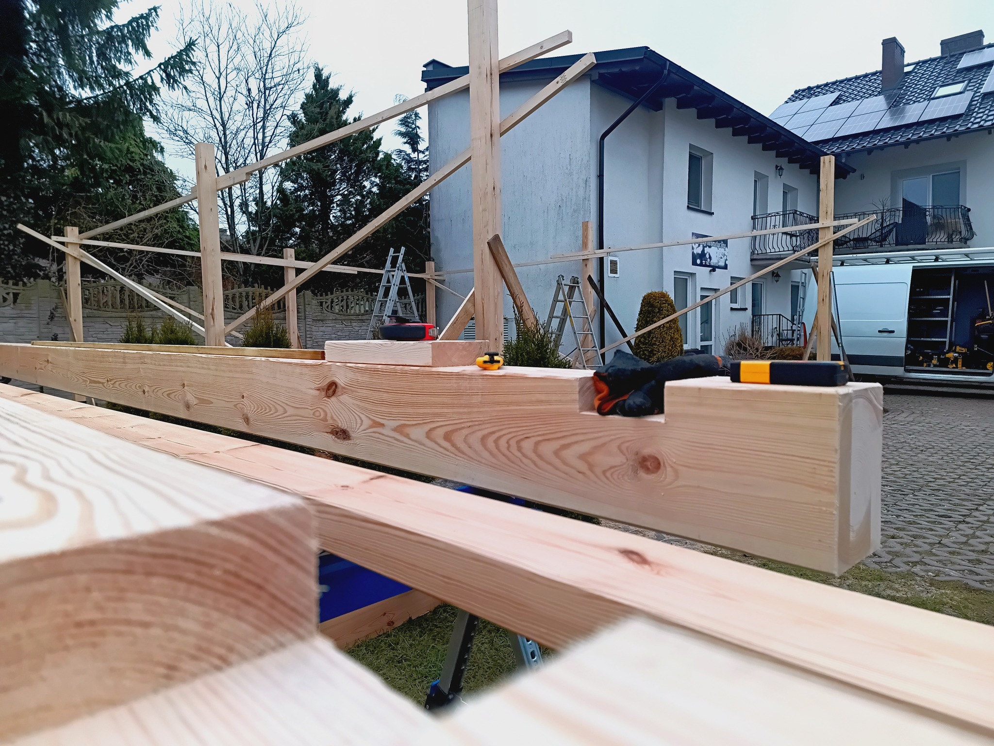 Wznoszenie drewnianej konstrukcji szkieletowej na tle domu z panelami słonecznymi. Widoczne narzędzia i drabiny, sugerujące etap budowy altany lub domu szkieletowego.