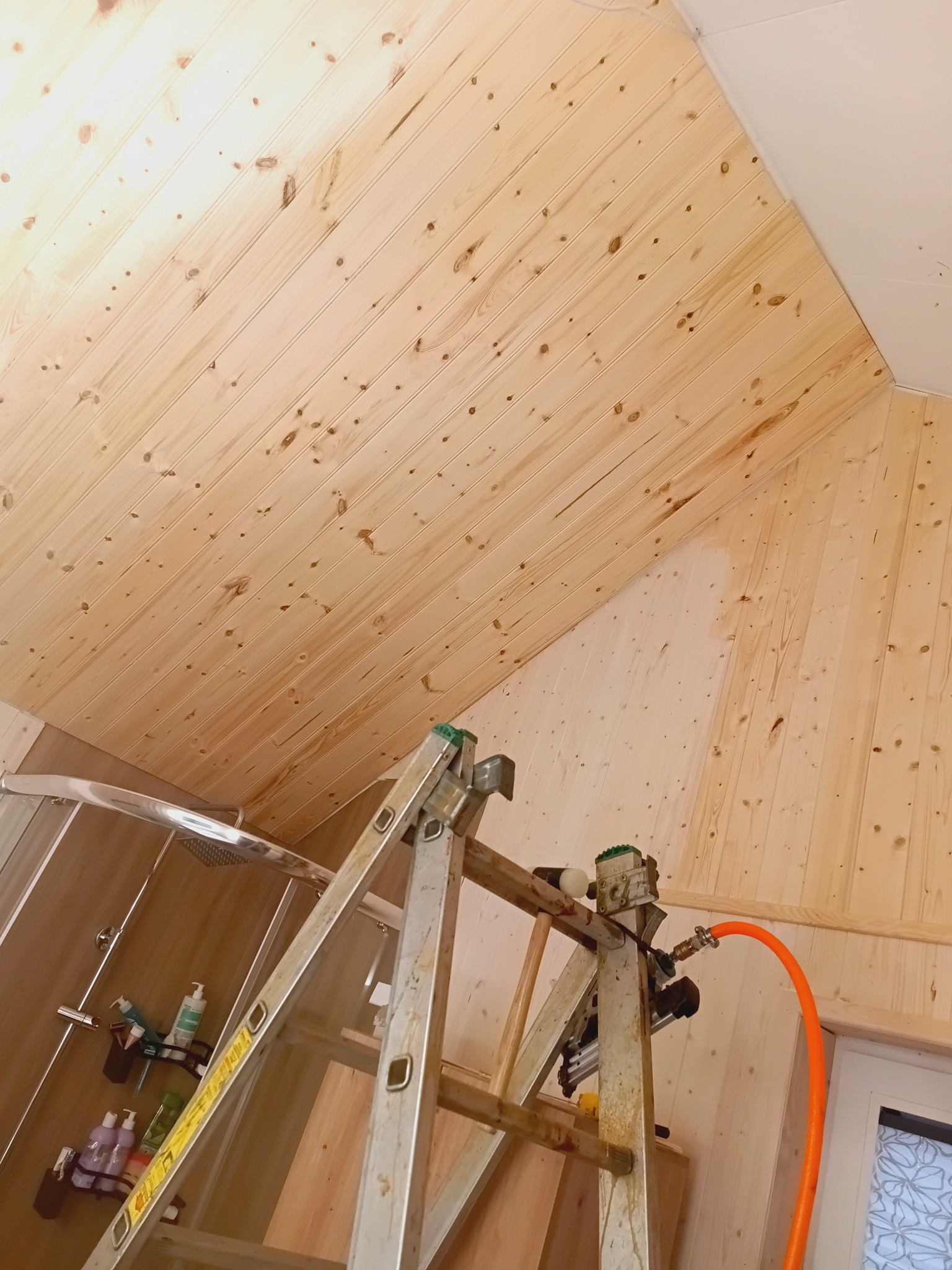 Wykończenie drewnem w łazience na poddaszu. Widoczna drabina z narzędziami do montażu boazerii. Ściany i sufit pokryte jasnym drewnem. Fragment kabiny prysznicowej.