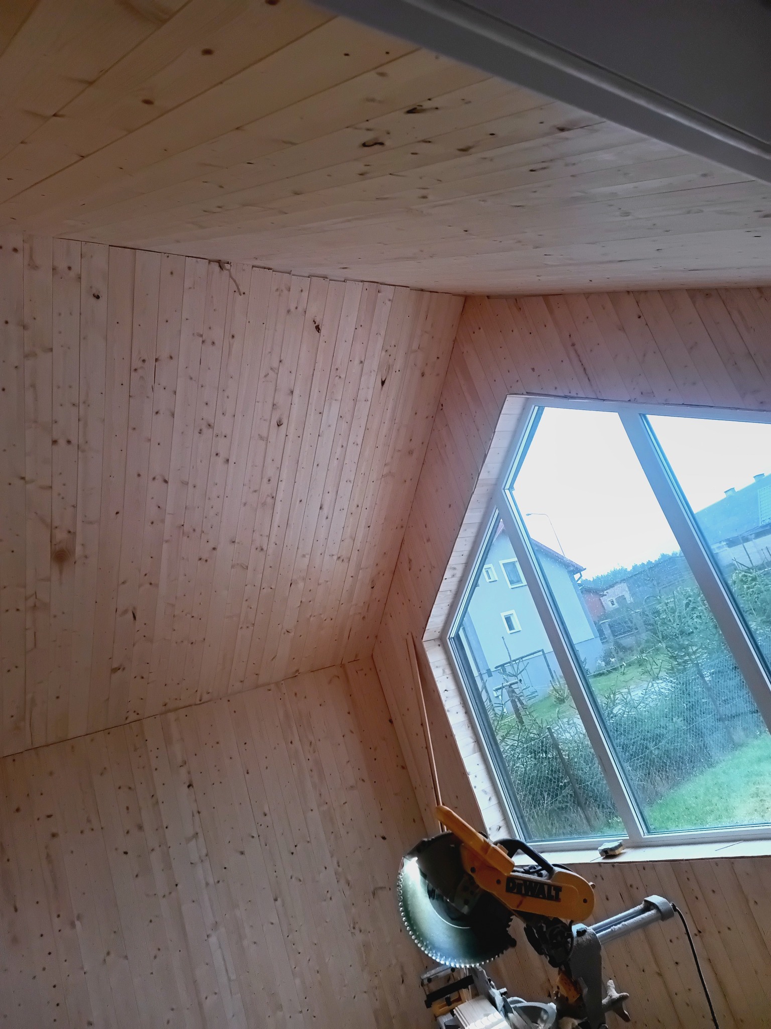 Wnętrze domu szkieletowego w trakcie wykańczania: drewniane panele ścienne i sufitowe, okno o nietypowym kształcie, piła ukośnica DeWalt. Widok z dołu.