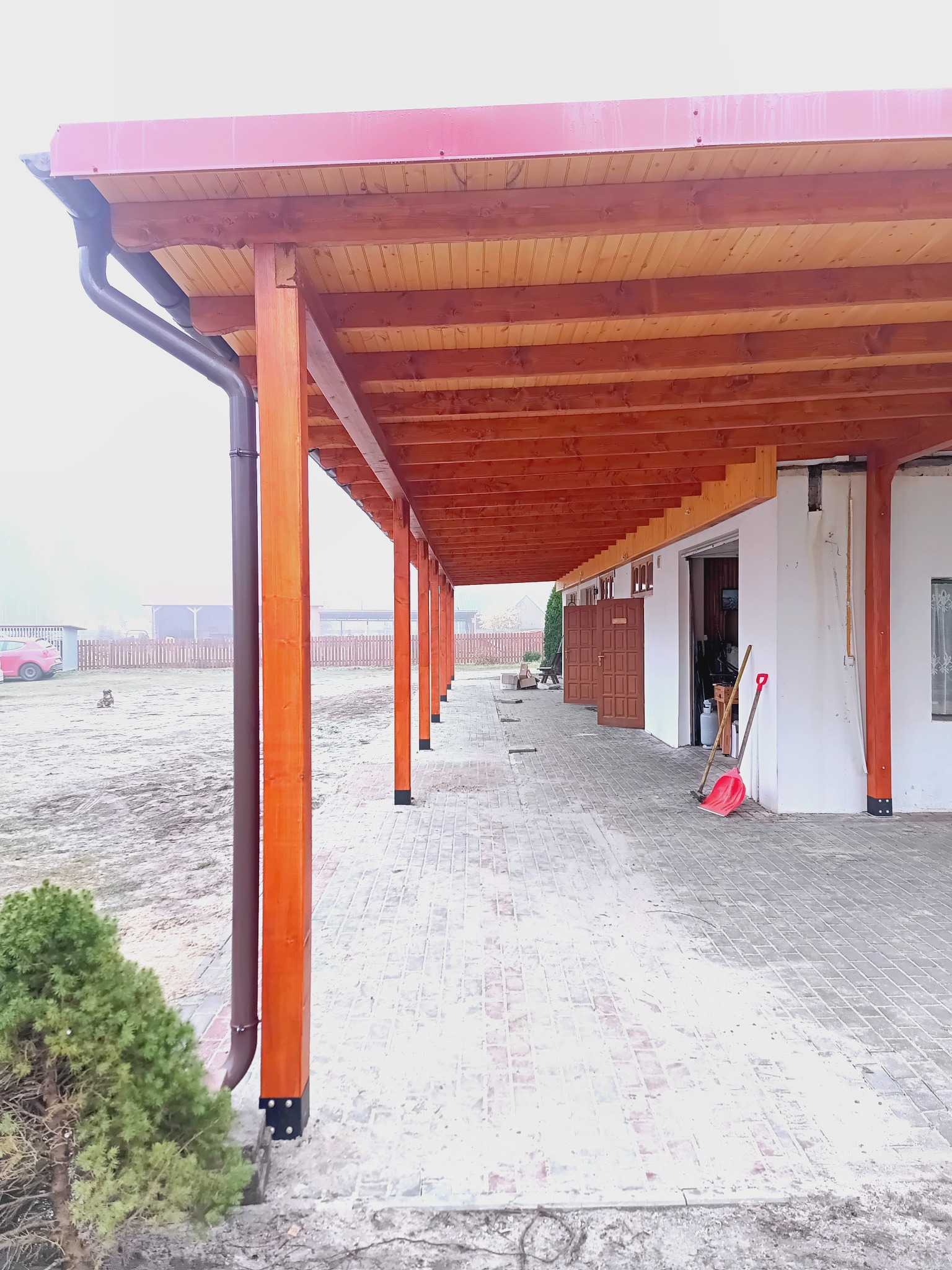 Zadaszenie tarasu z drewnianą konstrukcją, widok z perspektywy chodnika. Brązowe słupy, czerwona dachówka, widoczne rynny. W tle budynek z otwartymi drzwiami.