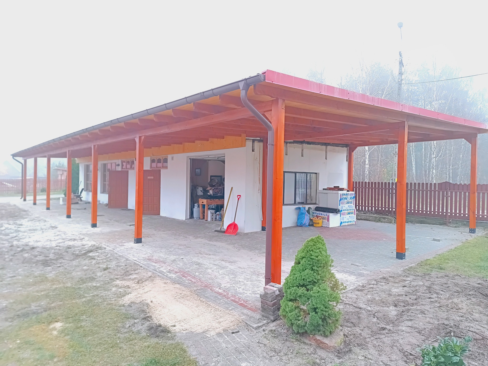 Zadaszenie tarasu z drewnianą konstrukcją i czerwonym dachem, widok z zewnątrz, kostka brukowa, elementy wykończeniowe, w tle budynek i drzewa.