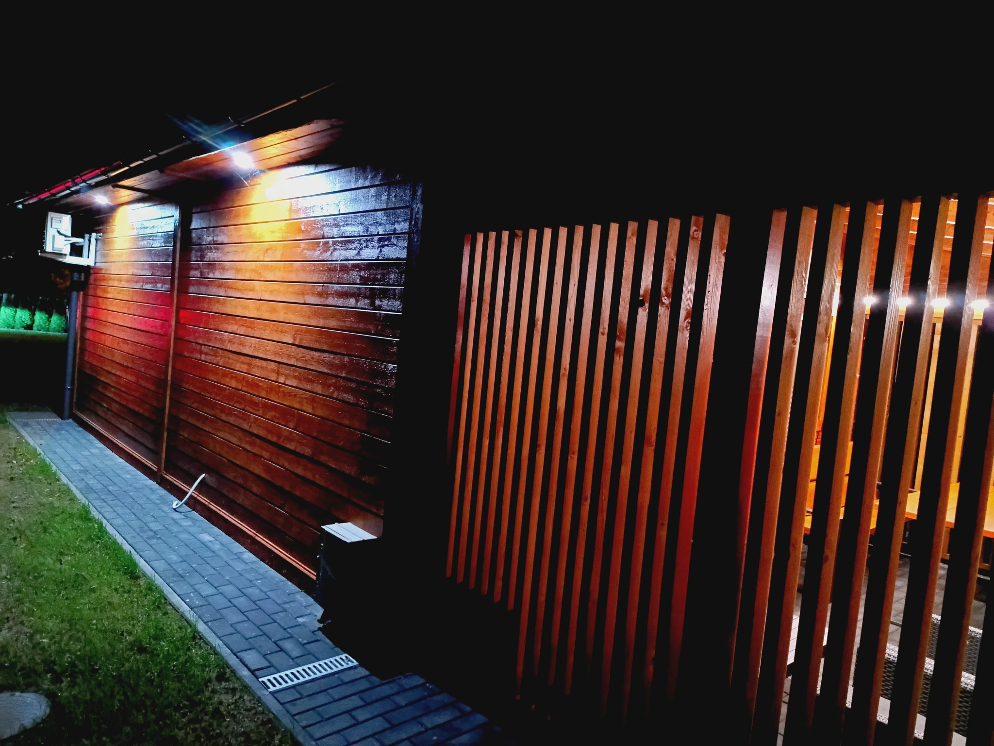 Nowoczesna, drewniana altana z oświetleniem nocnym, widoczna z zewnątrz przez ażurową ścianę. Kostka brukowa i trawnik dopełniają otoczenie.