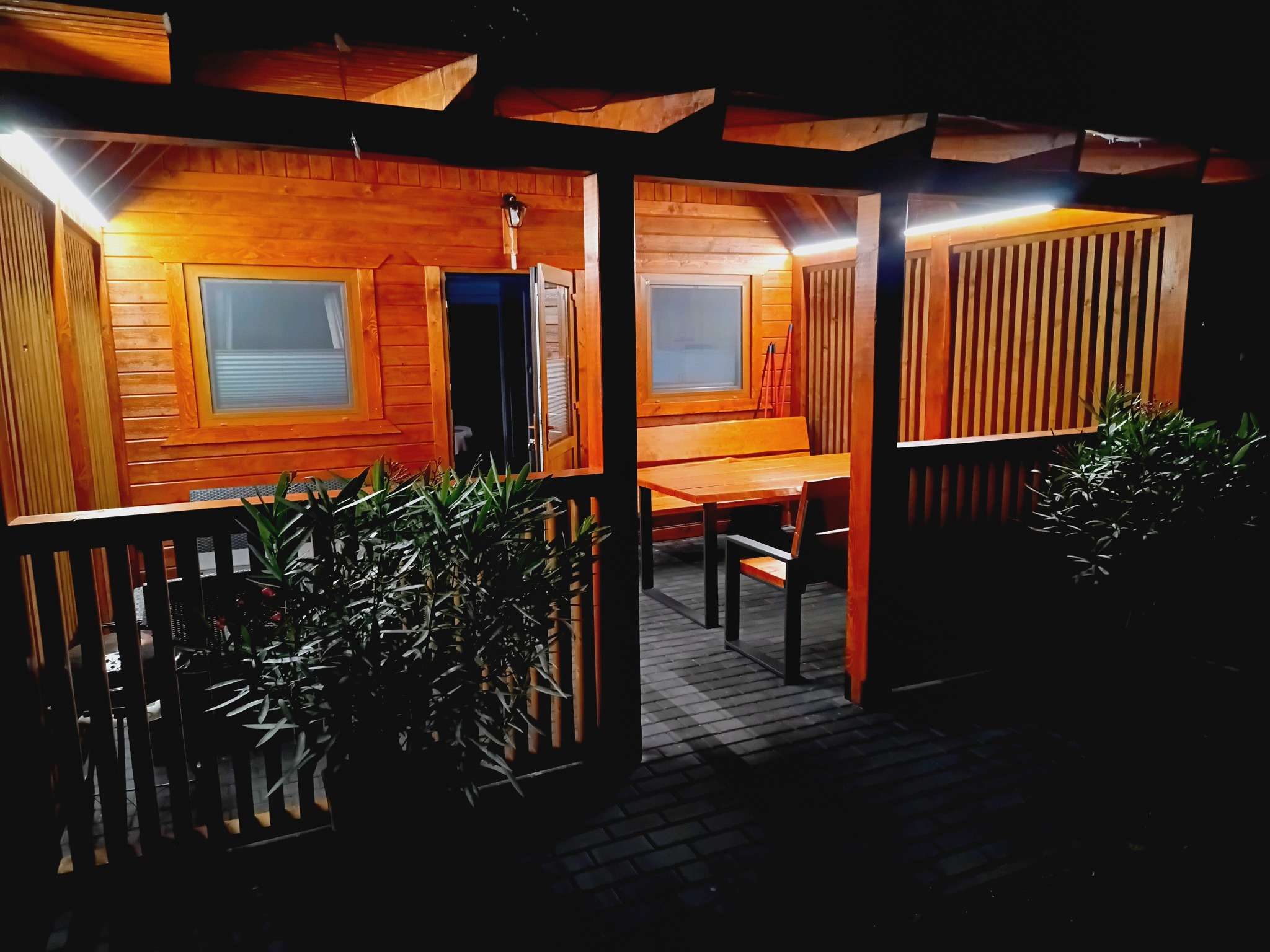 Oświetlona altana z drewna z oknami i meblami ogrodowymi, widok nocą. Rośliny w donicach na pierwszym planie, ciepłe światło podkreśla drewnianą konstrukcję.