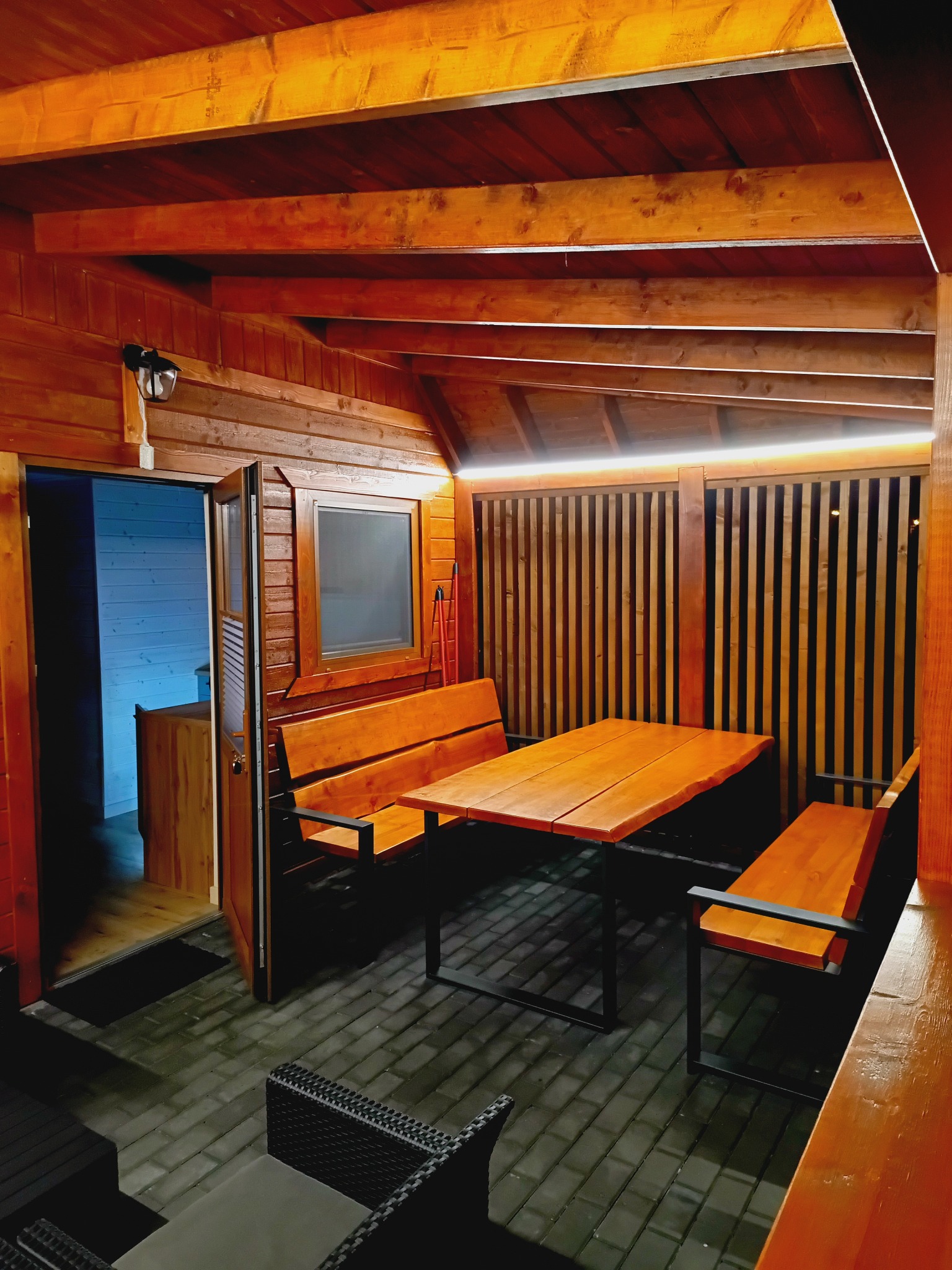 Drewniana altana z oświetleniem LED, wyposażona w stół i ławki. Widoczne wejście do pomieszczenia z oknem. Podłoga wyłożona kostką brukową.