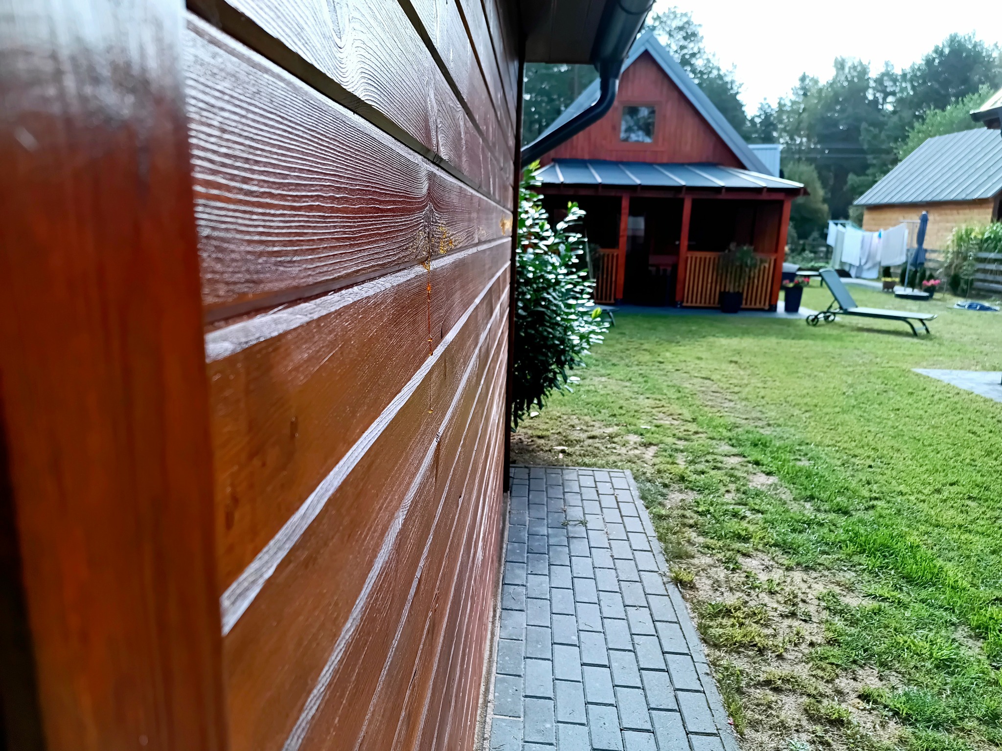 Ściana domu szkieletowego z drewnianą elewacją, widoczna z bliska, z kroplą żywicy. W tle trawnik i drugi budynek z werandą.