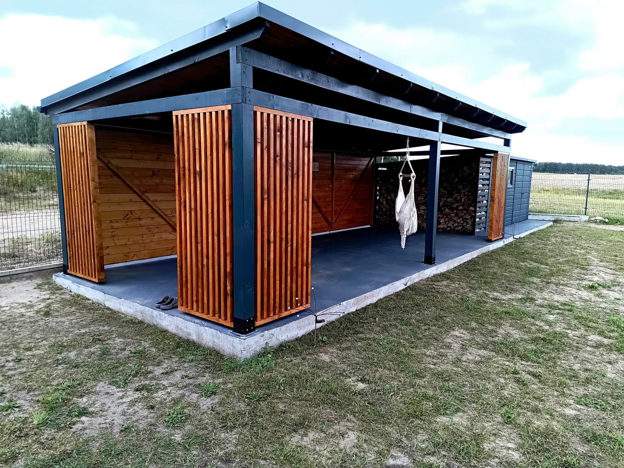 Nowoczesna altana z drewna i metalu, z elementami dekoracyjnymi z listewek. Wewnątrz widoczny hamak i drewno na opał. Całość na betonowej wylewce, otoczona trawnikiem.