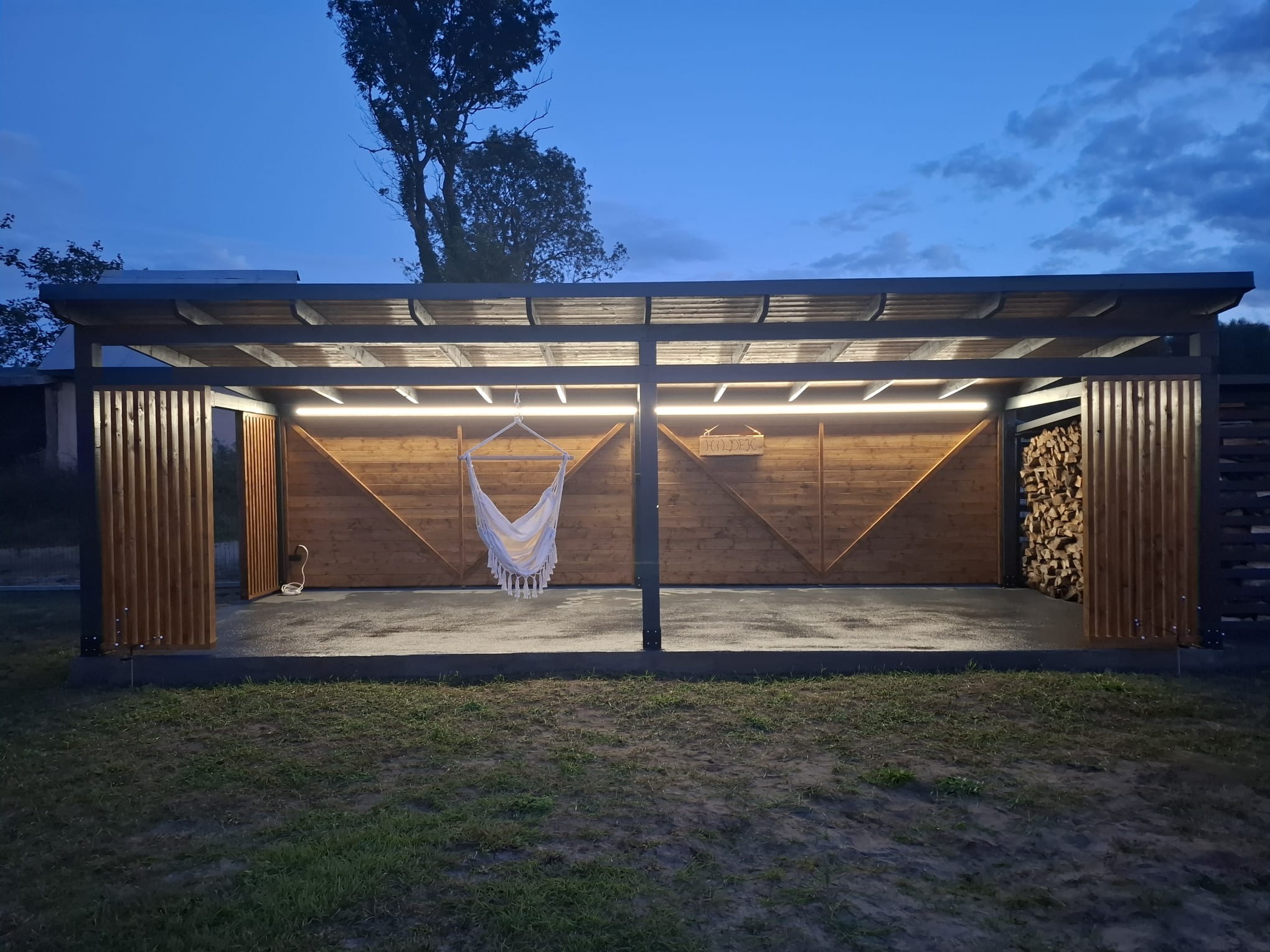Nowoczesna altana z drewna i metalu, oświetlona ledami, z hamakiem i zapasem drewna. Ujęcie wieczorne, podkreślające ciepłe światło i przytulny charakter.