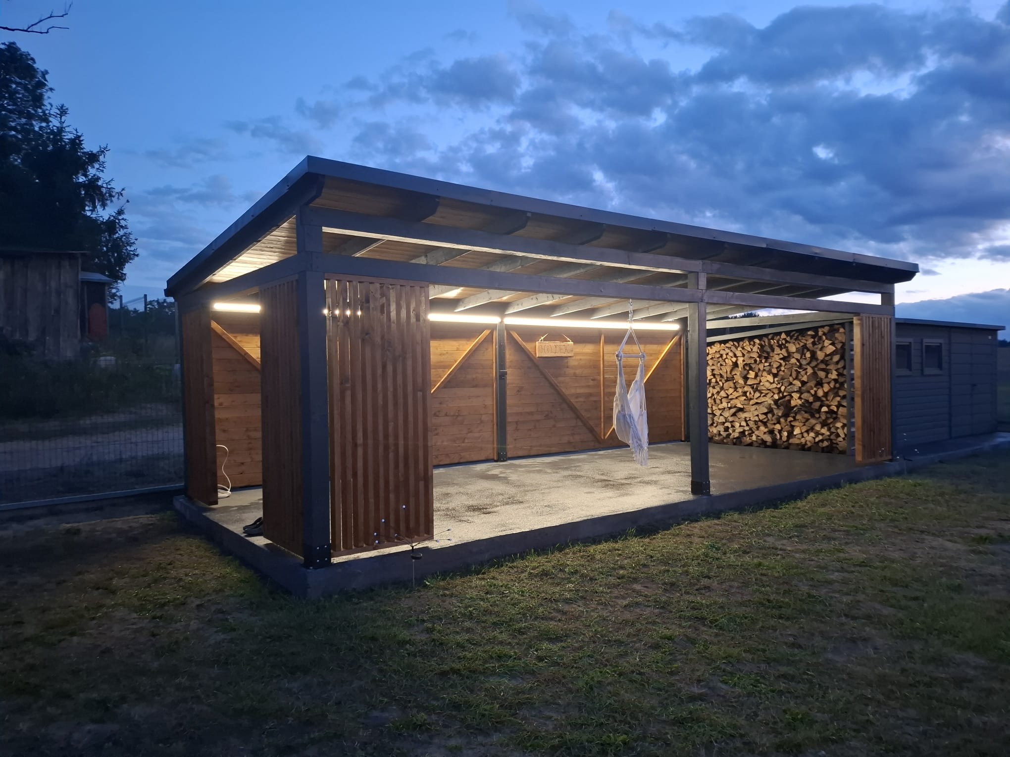 Nowoczesna altana z drewna z oświetleniem LED, częściowo zabudowana, z miejscem na drewno. Widok wieczorny, z zewnątrz, na tle zachmurzonego nieba.