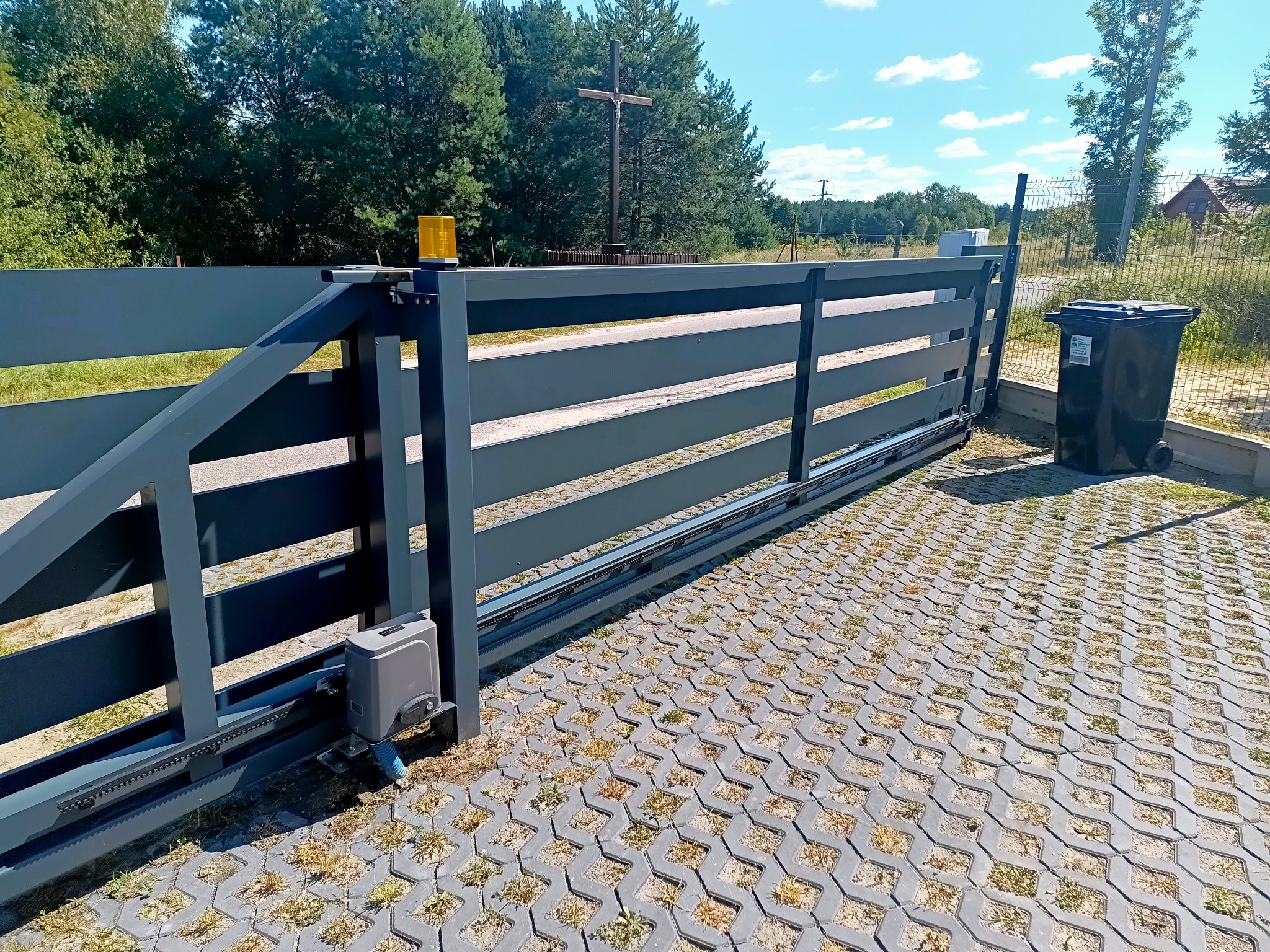 Szara, przesuwna brama wjazdowa z automatyką, widoczna z boku na tle zieleni i krzyża. Kostka brukowa z trawą w szczelinach. Żółta lampa ostrzegawcza na bramie.