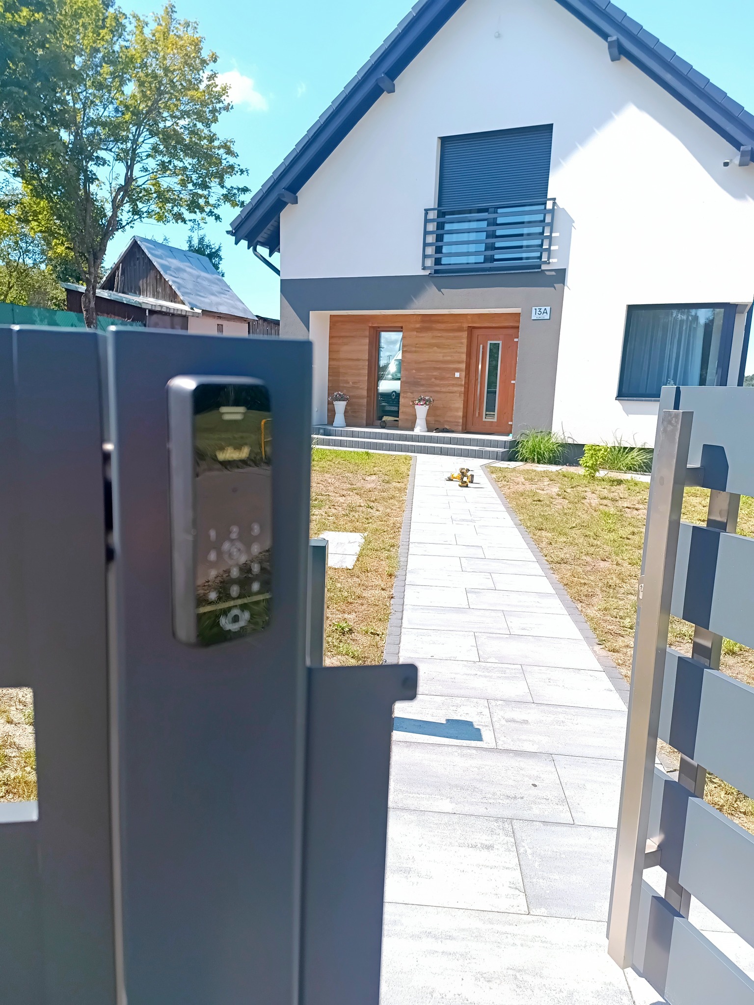 Nowoczesny dom jednorodzinny z szarą bramą z panelem dotykowym. Widok na ścieżkę z kostki brukowej i elewację z drewnianymi akcentami. Słoneczny dzień.