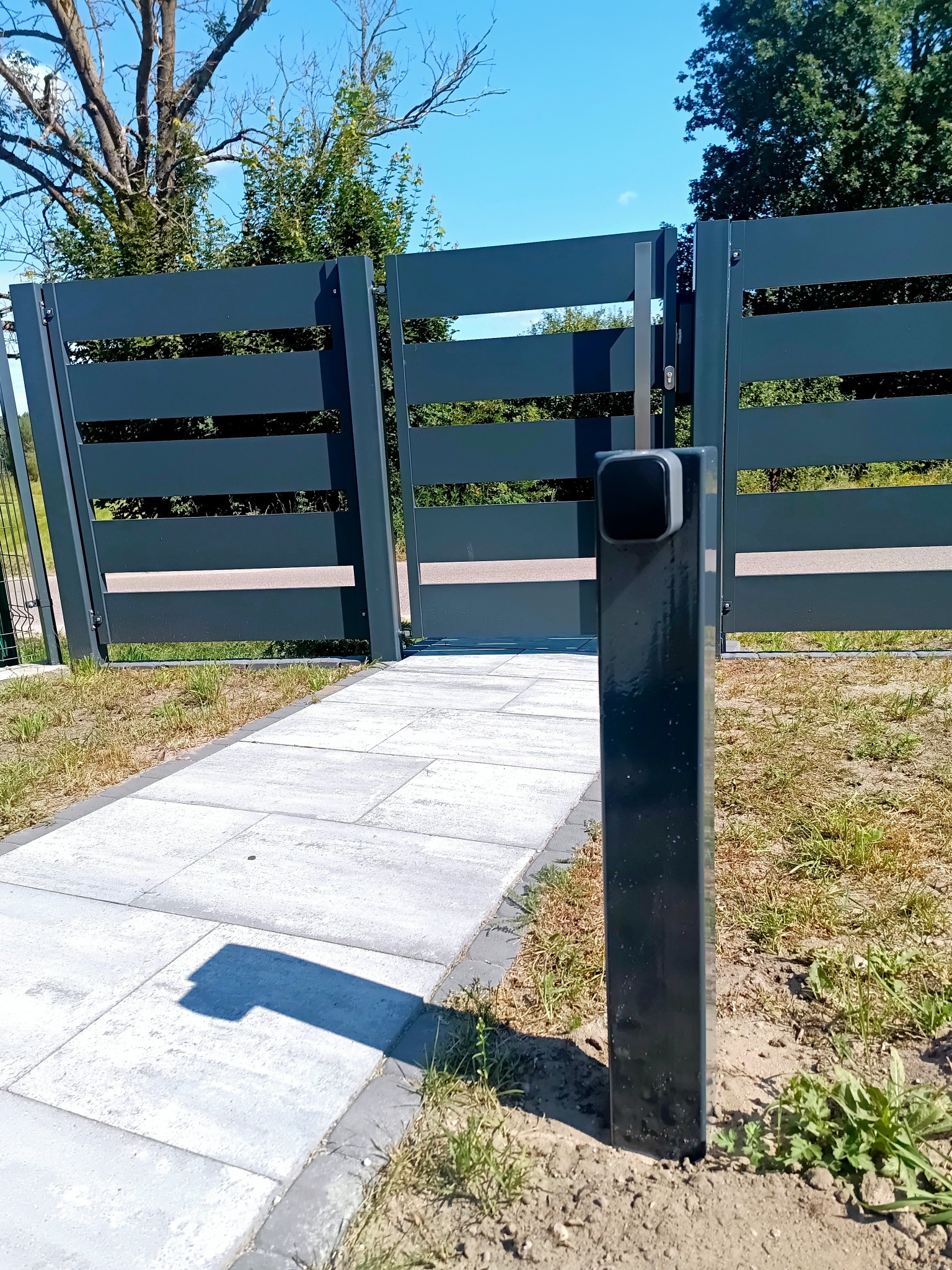 Nowoczesna, grafitowa brama przesuwna z poziomymi panelami, otwierająca się na wybrukowaną ścieżkę w słoneczny dzień. Widoczny słupek z domofonem.