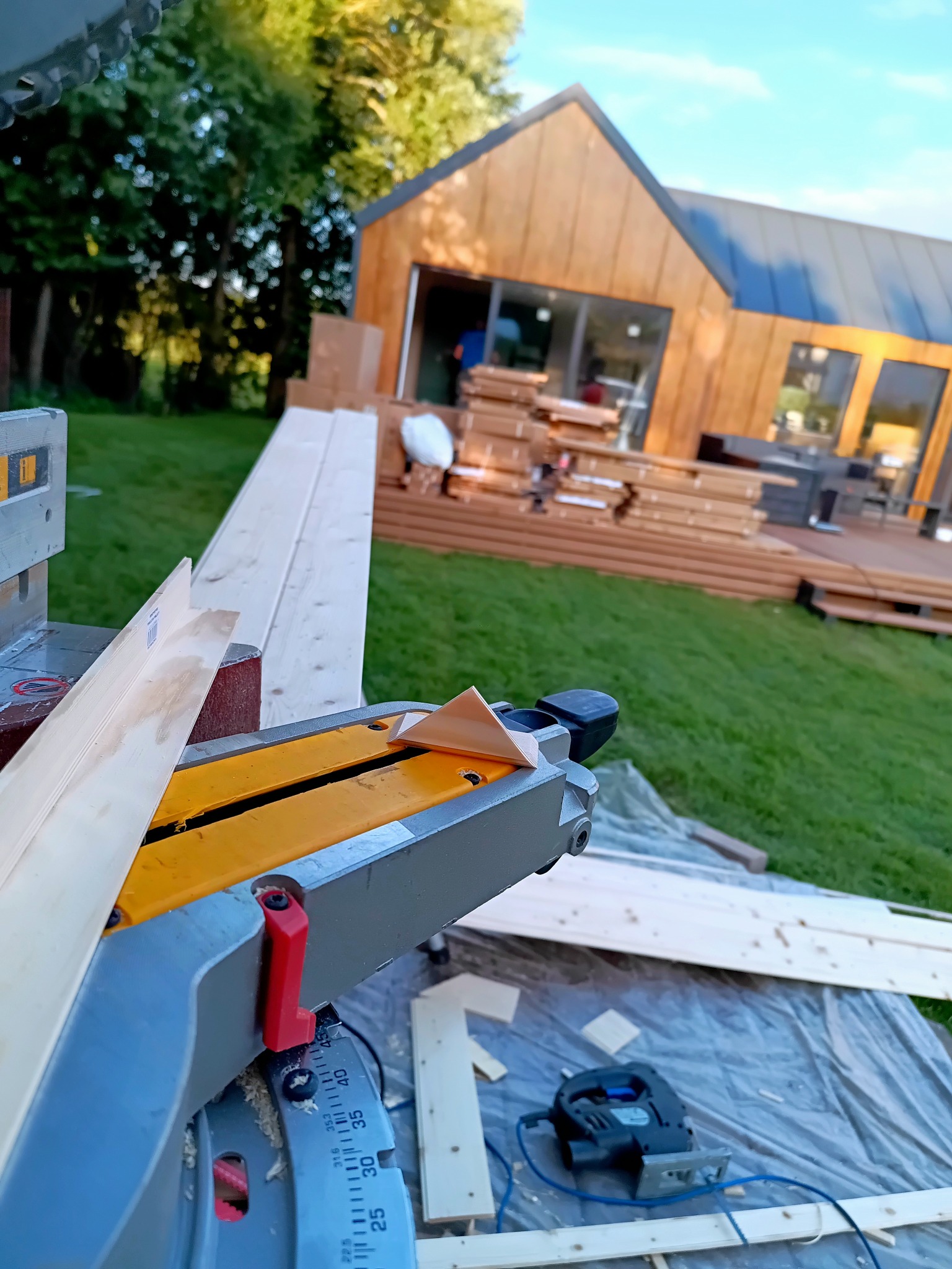 Budowa domu szkieletowego: piła ukośnica tnie deskę na tle drewnianego domu z tarasem i stertą desek. Widoczne narzędzia i elementy konstrukcyjne.