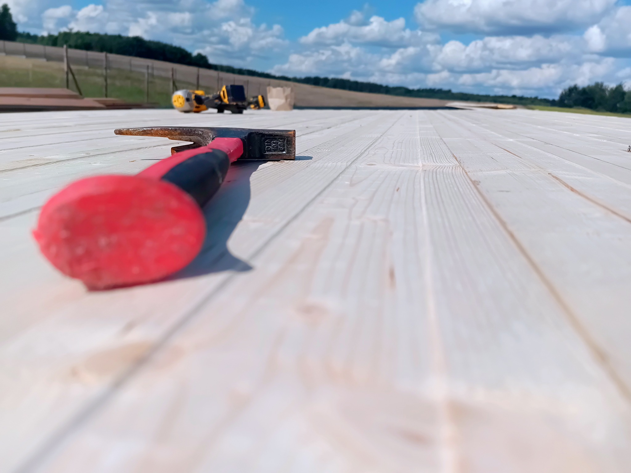 Młotek z czerwoną rączką leży na drewnianej podłodze w trakcie budowy domu szkieletowego. W tle widoczne maszyny budowlane i błękitne niebo.