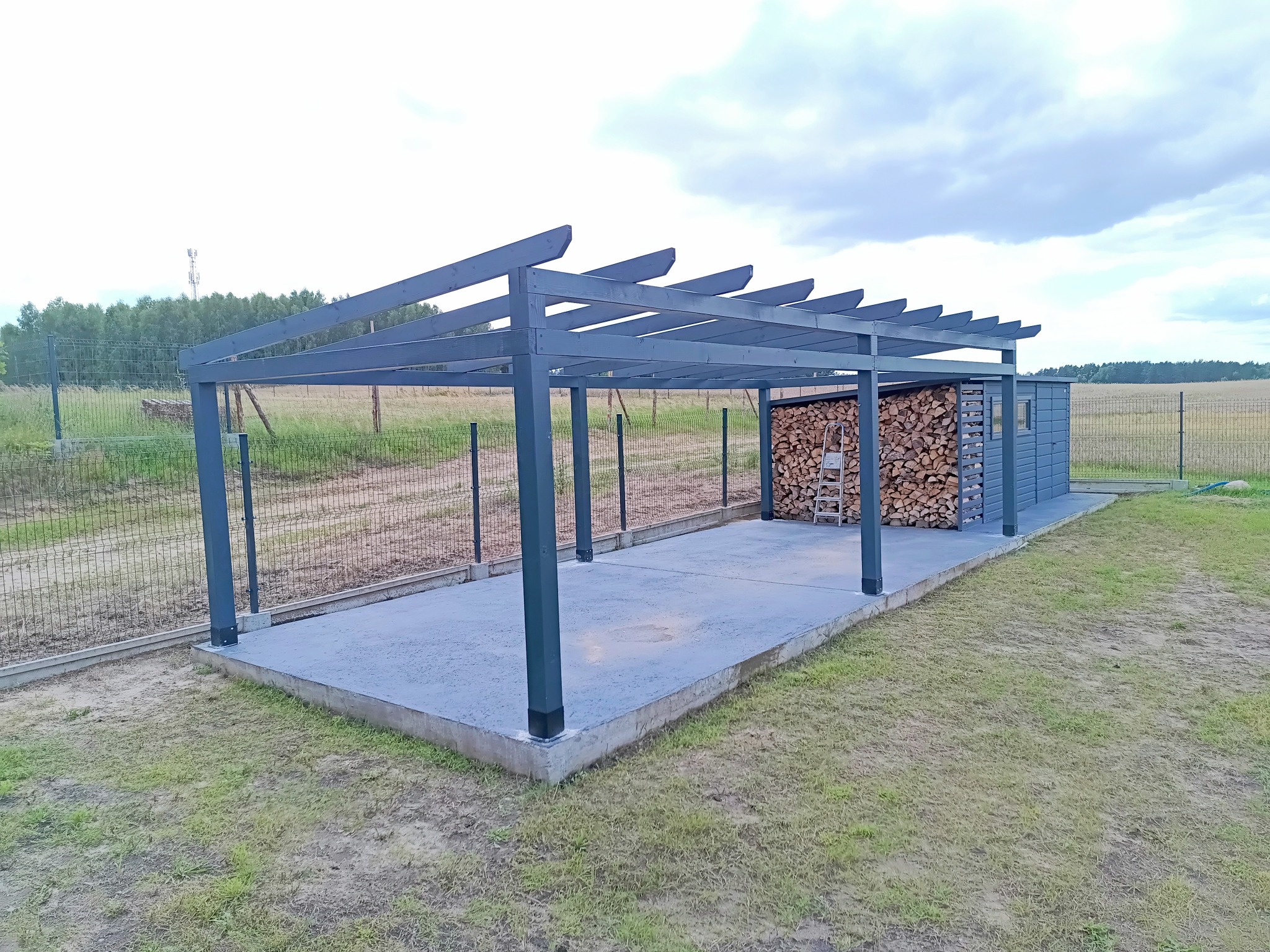 Szara, drewniana altana z zadaszeniem lamelowym i drewutnią, na betonowej wylewce, otoczona siatką ogrodzeniową i zielenią. Widok z perspektywy.