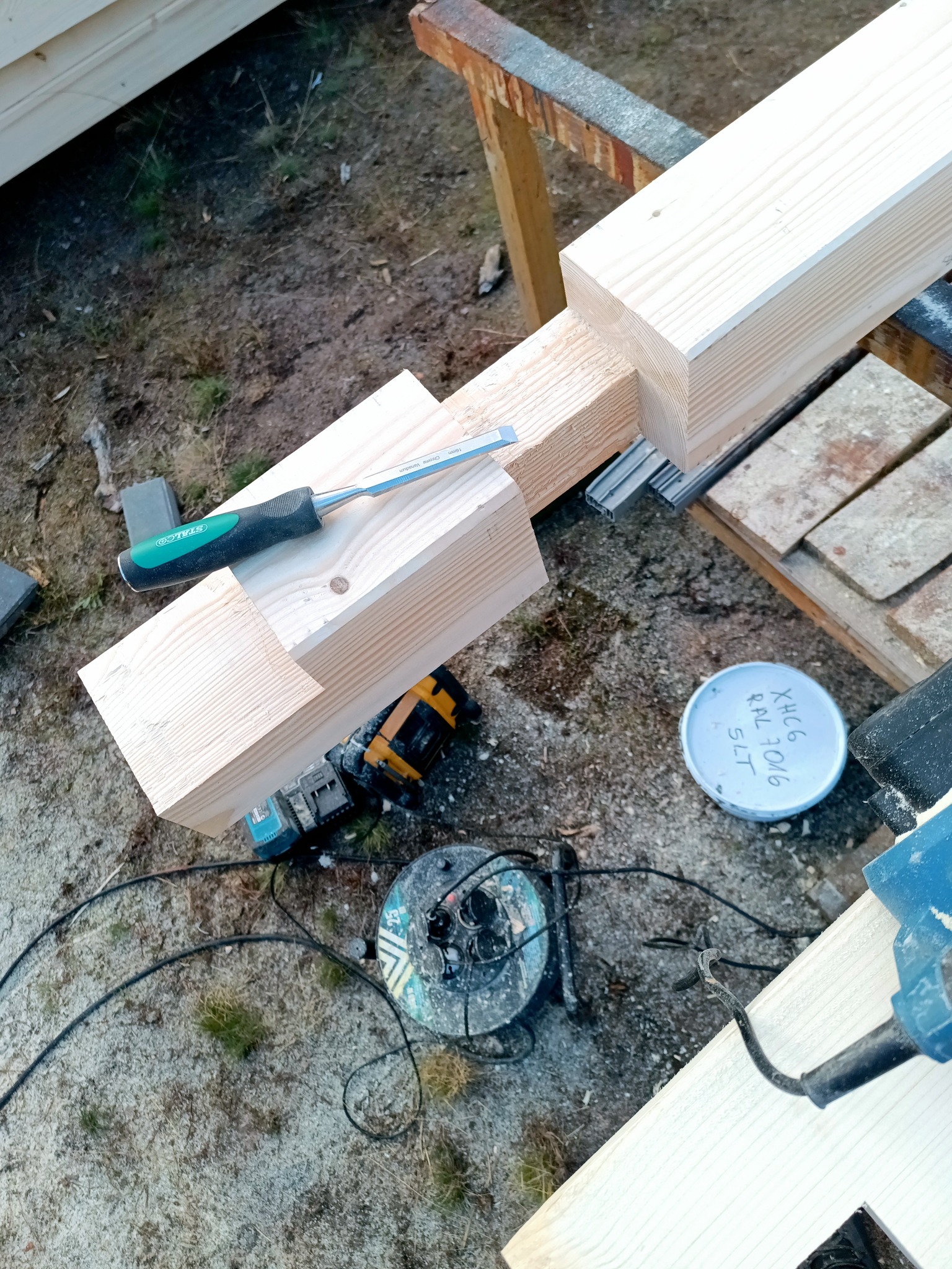 Fragment konstrukcji drewnianej z dłutem, wiertarką i puszką farby. Detal łączenia elementów, narzędzia pracy na ziemi. Budowa altany lub domu szkieletowego.